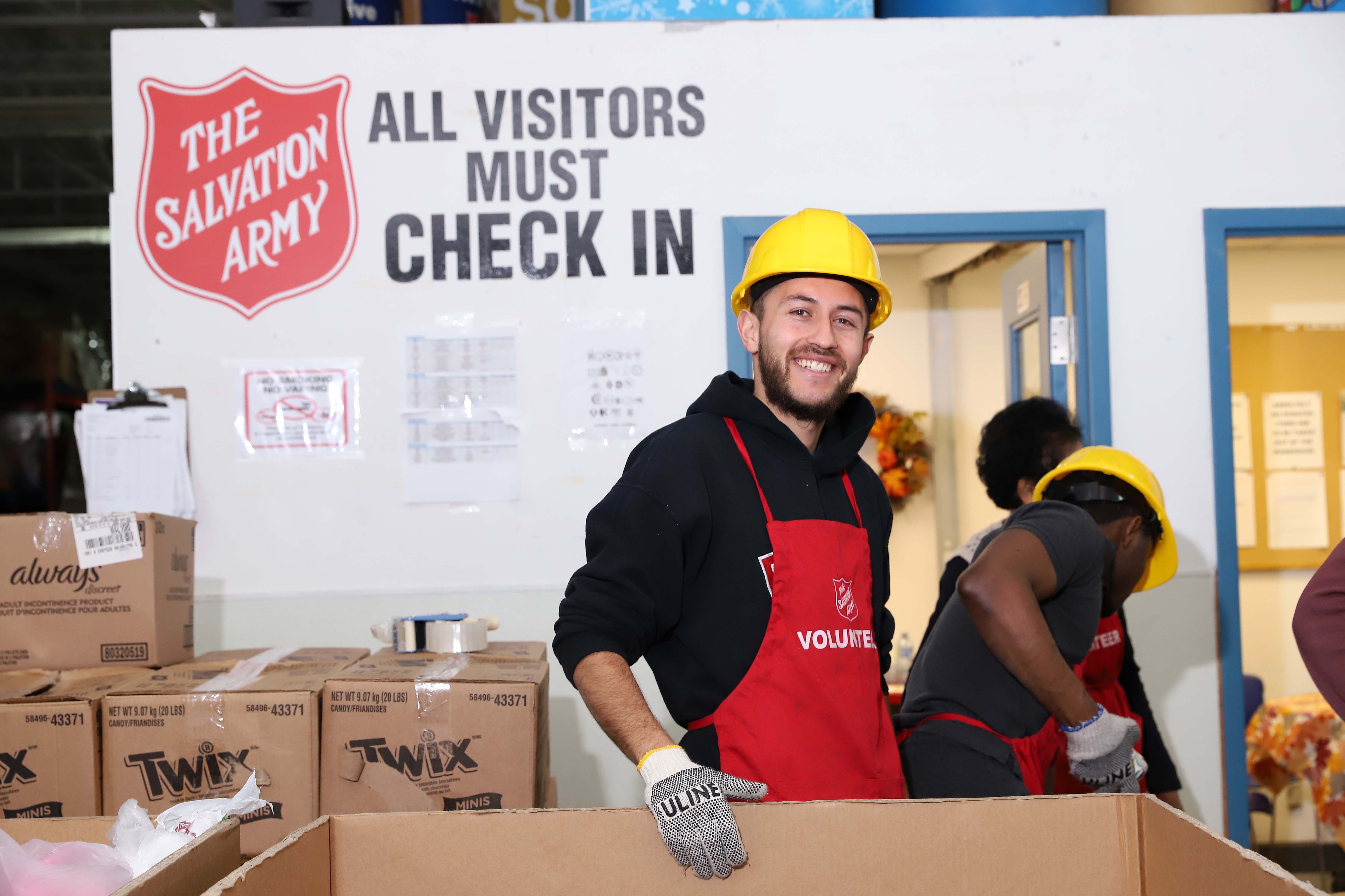 A smiling food bank volunteer in a warehouse, wearing a hard hat and gloves.