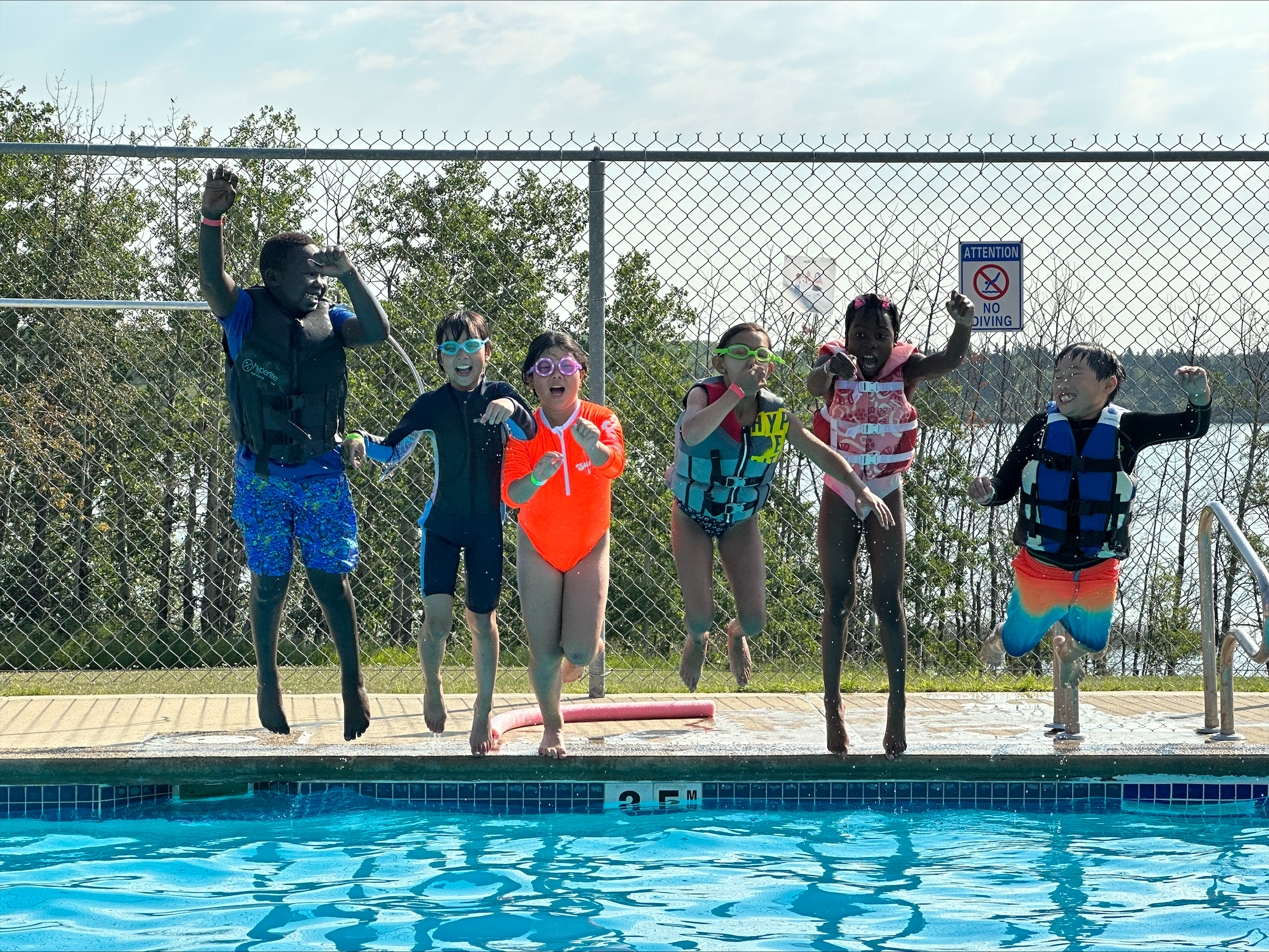 A picture of six kids in swimsuits, goggles and swimsuits jumping into a pool.