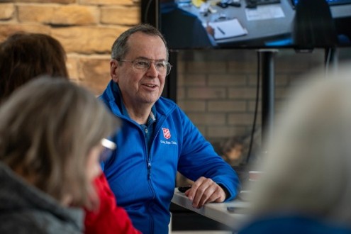 A Salvation Army mission partner wearing a blue Army sweater, speaking with a group of others at a board table. 